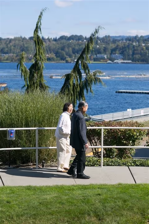 An elderly man and woman walking along a paved path with a metal railing near a body of water. There are green bushes and grass around them, and trees and hills are visible across the water under a clear sky.