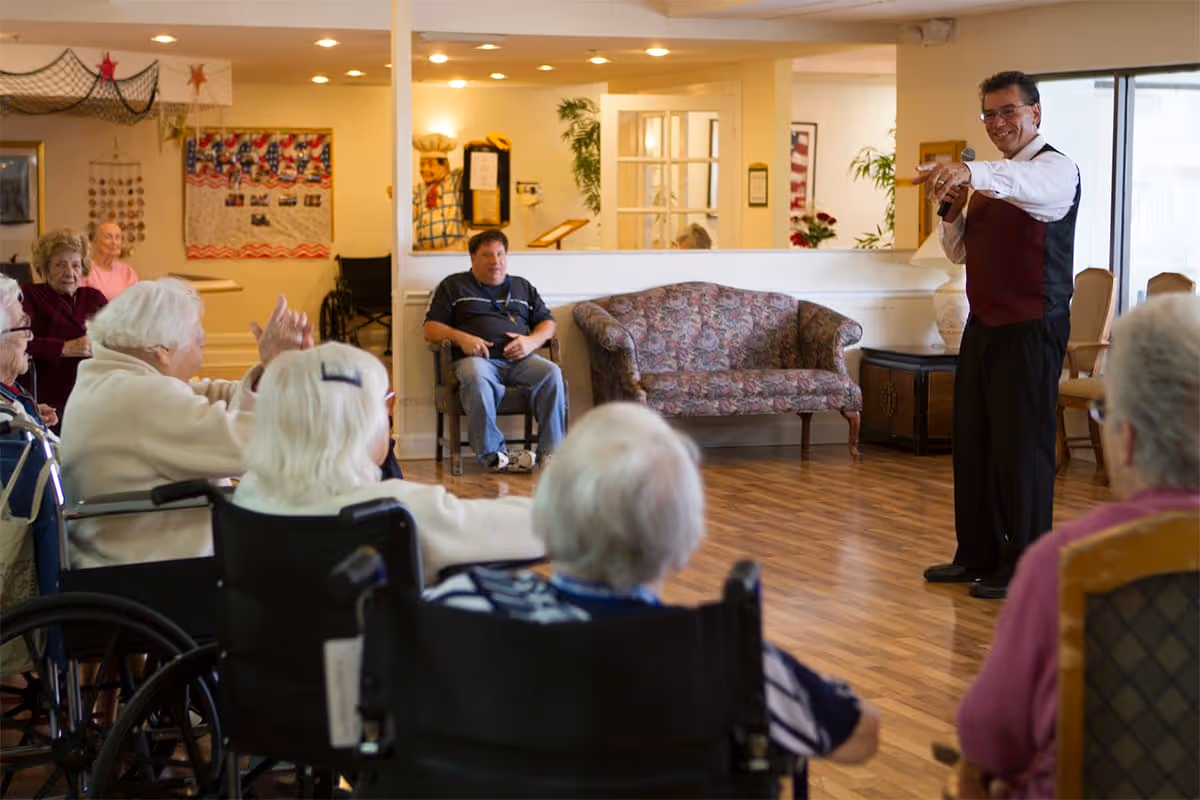 A group of elderly people seated in wheelchairs and chairs in a common room, attentively listening to a man holding a microphone and pointing, who appears to be leading an activity or event. The room has wooden flooring, a floral-patterned couch, and light-colored walls with decorations.