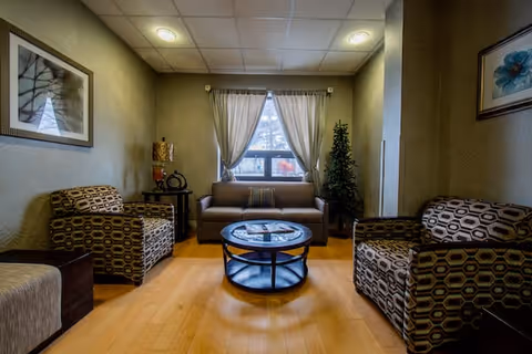 A cozy living room with a brown sofa centered against a window with white curtains. Two patterned armchairs are positioned on either side of a round glass coffee table. The room has wooden flooring, a side table with a lamp, framed artwork on the walls, and a tall plant in the corner.