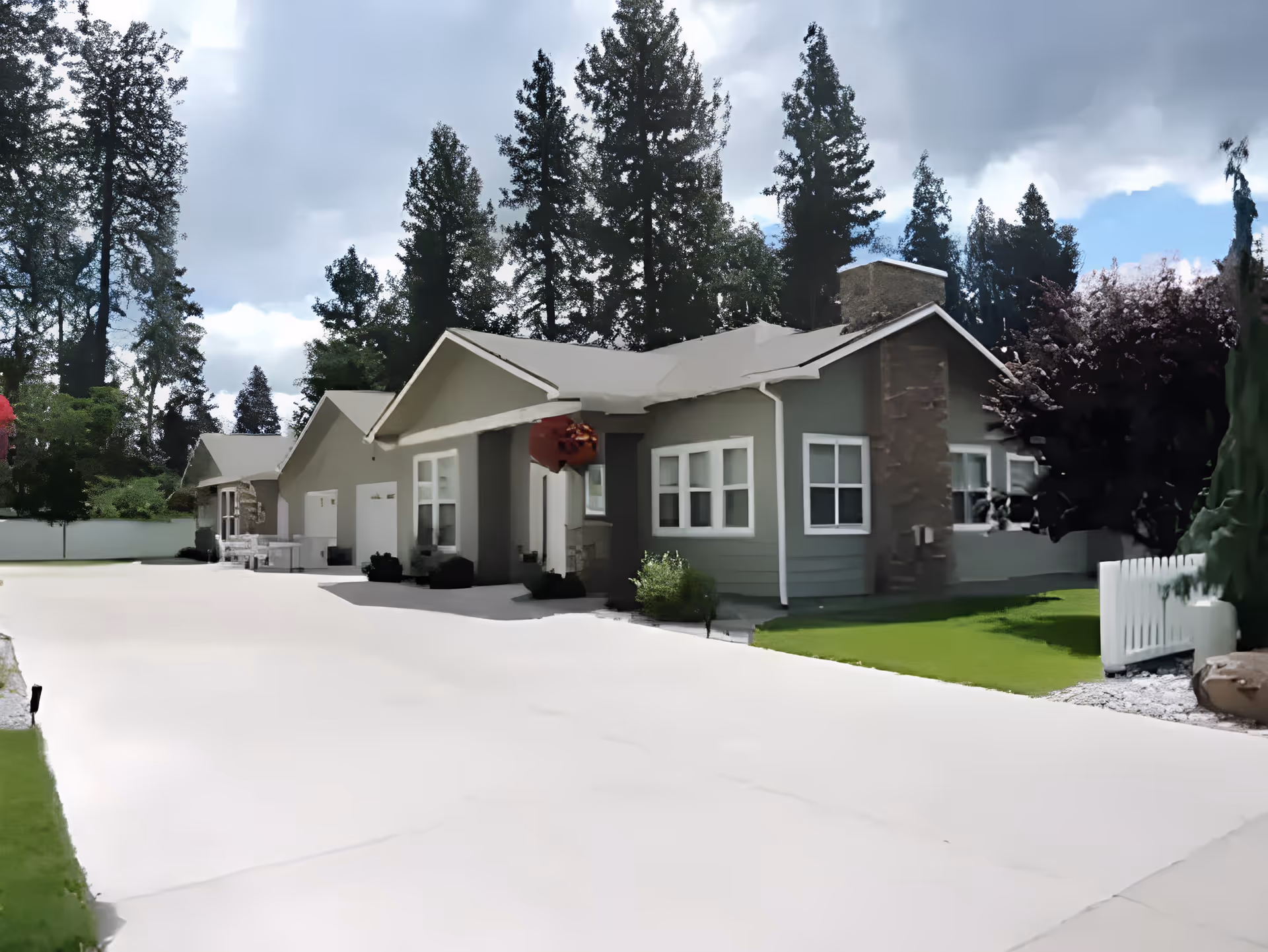 Exterior view of a single-story residential building with a driveway, green lawn, and tall trees in the background under a cloudy sky.