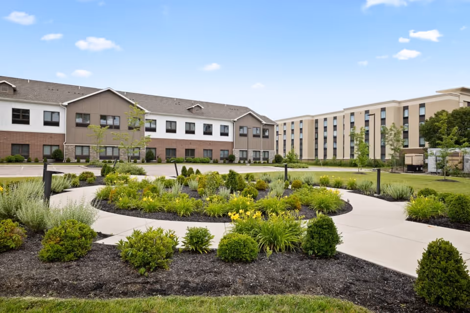Landscaped circular walkway and garden in front of a multi-story senior living facility building under a blue sky.