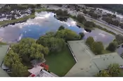 Aerial view of a senior living facility named Highlands Lake Center, showing multiple buildings with green roofs surrounding a large pond with reflections of clouds and sky. There are trees and landscaped green areas around the buildings and water.