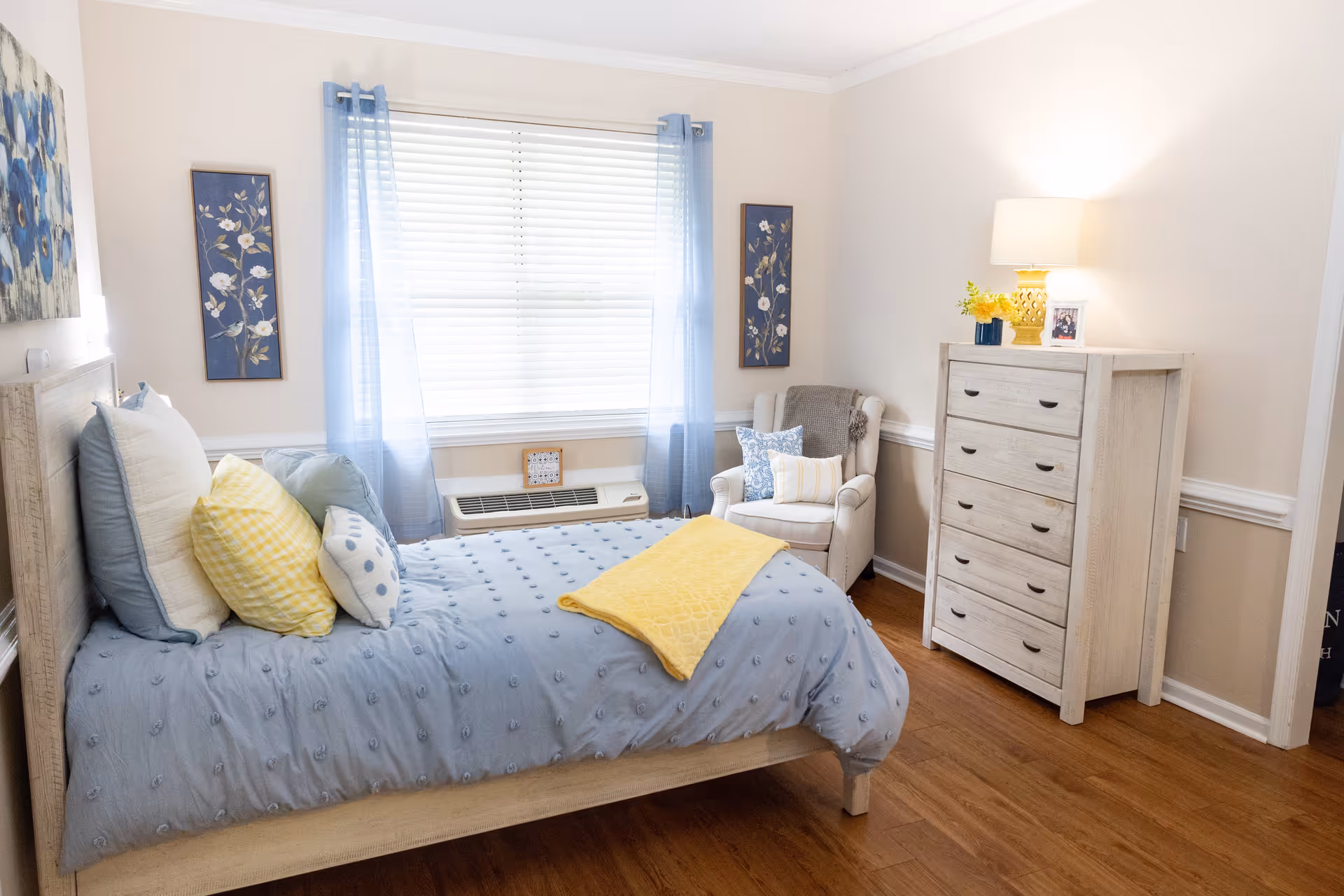 Sunlit bedroom with a bed dressed in blue and yellow linens, an armchair by the window, and a light wood dresser.