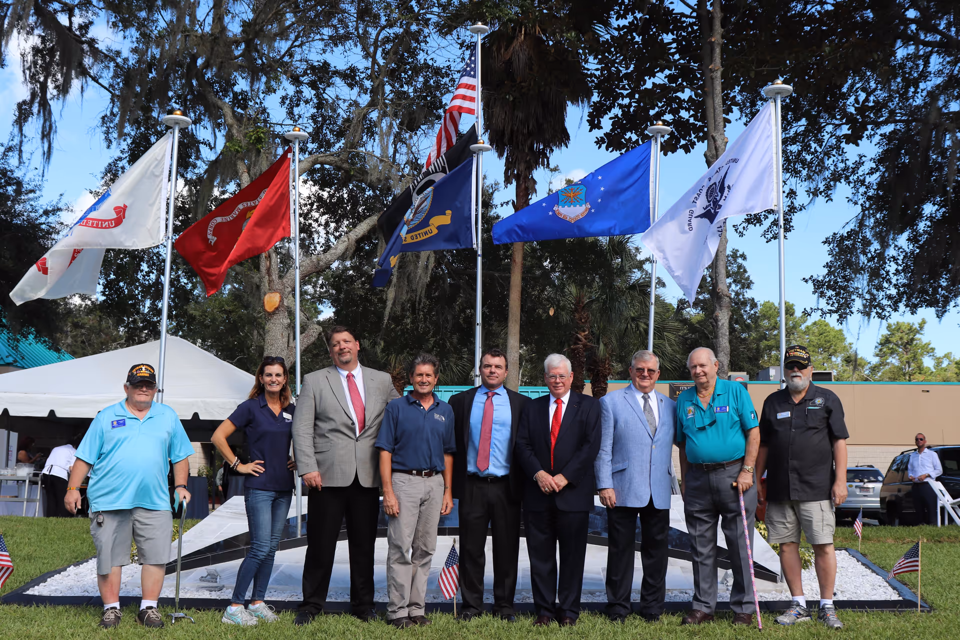 A group of nine people standing outdoors in front of several flagpoles displaying various flags, including the American flag. The group includes men and women dressed in casual and formal attire, with trees and a tent in the background.