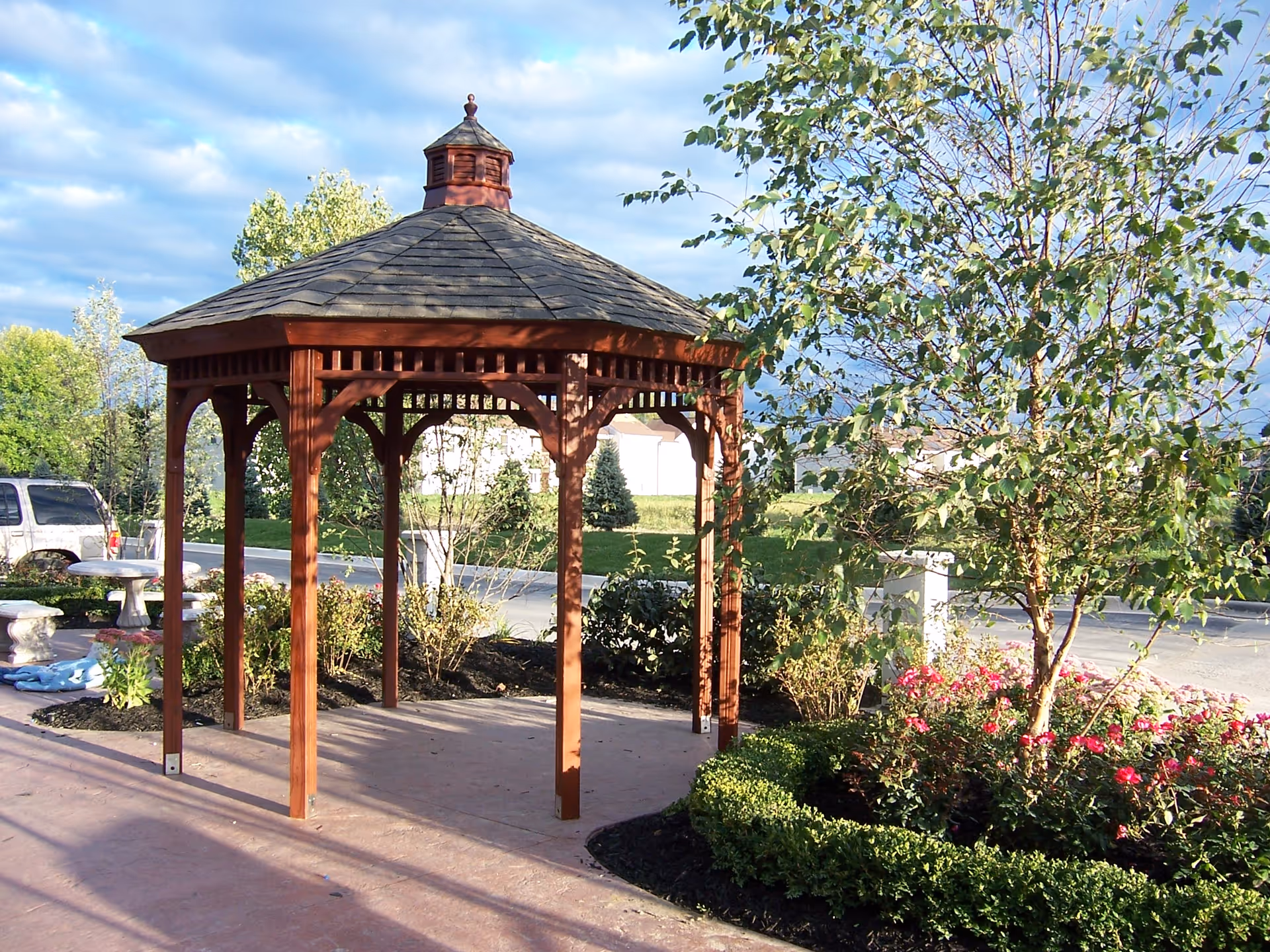 A wooden gazebo with a shingled roof situated on a paved area surrounded by landscaped greenery including bushes, flowers, and trees under a partly cloudy sky.