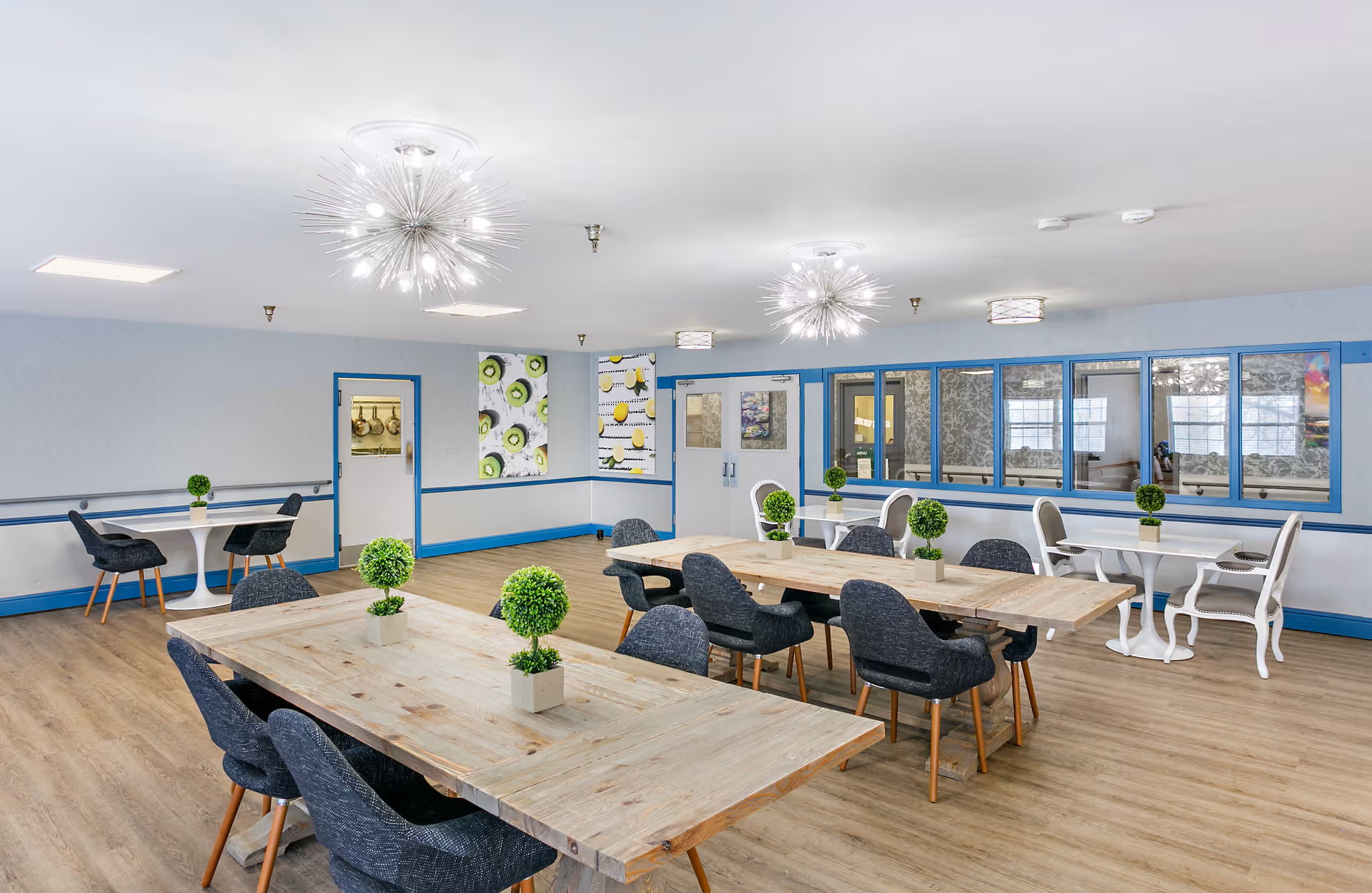 Bright communal dining/activity room with wooden tables, gray upholstered chairs, decorative pendant lights, and small potted plants.