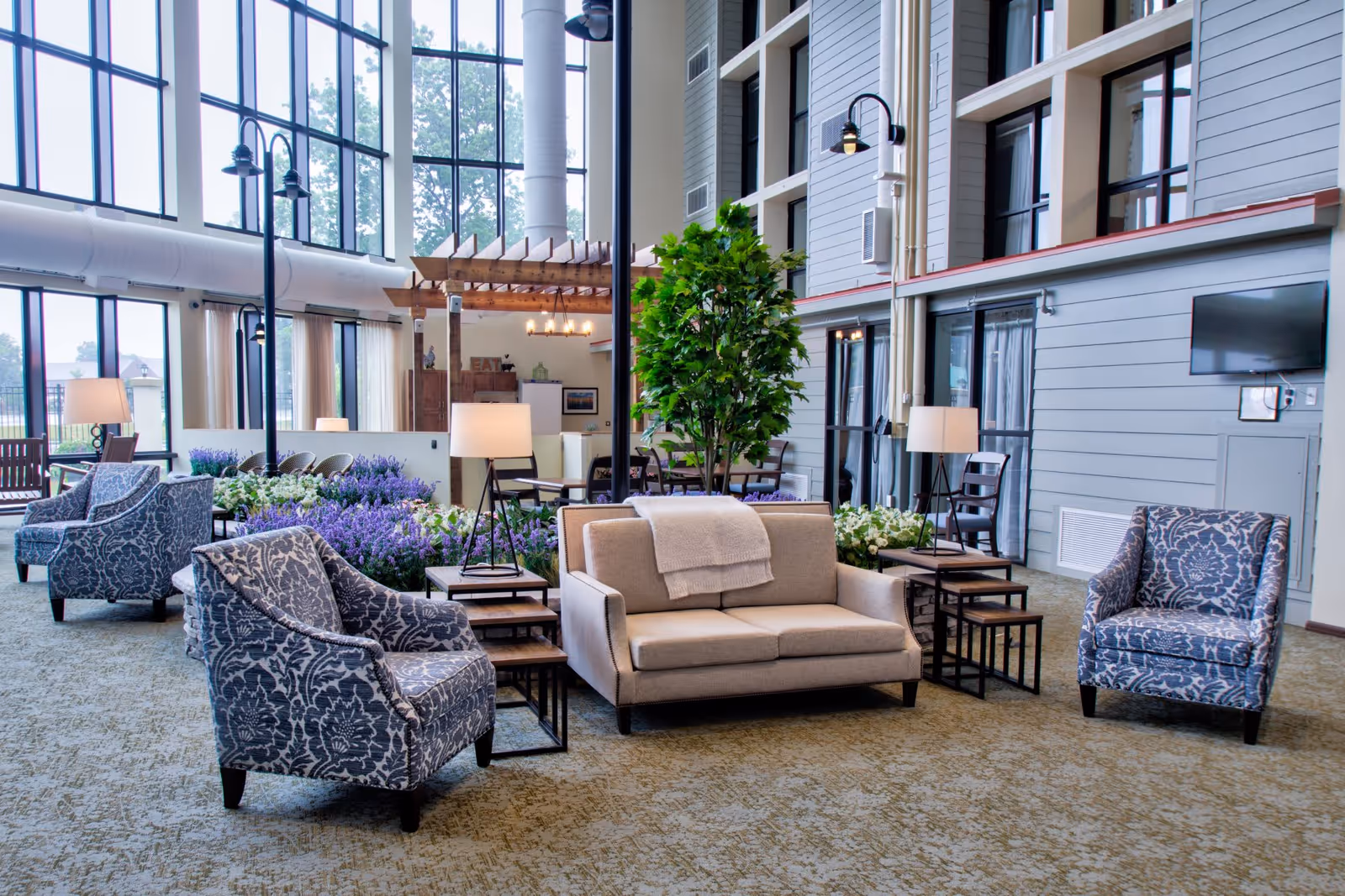 A spacious and well-lit living area in a senior living facility with large floor-to-ceiling windows, patterned armchairs, a beige sofa with a throw blanket, side tables with lamps, indoor plants, and a wall-mounted TV.