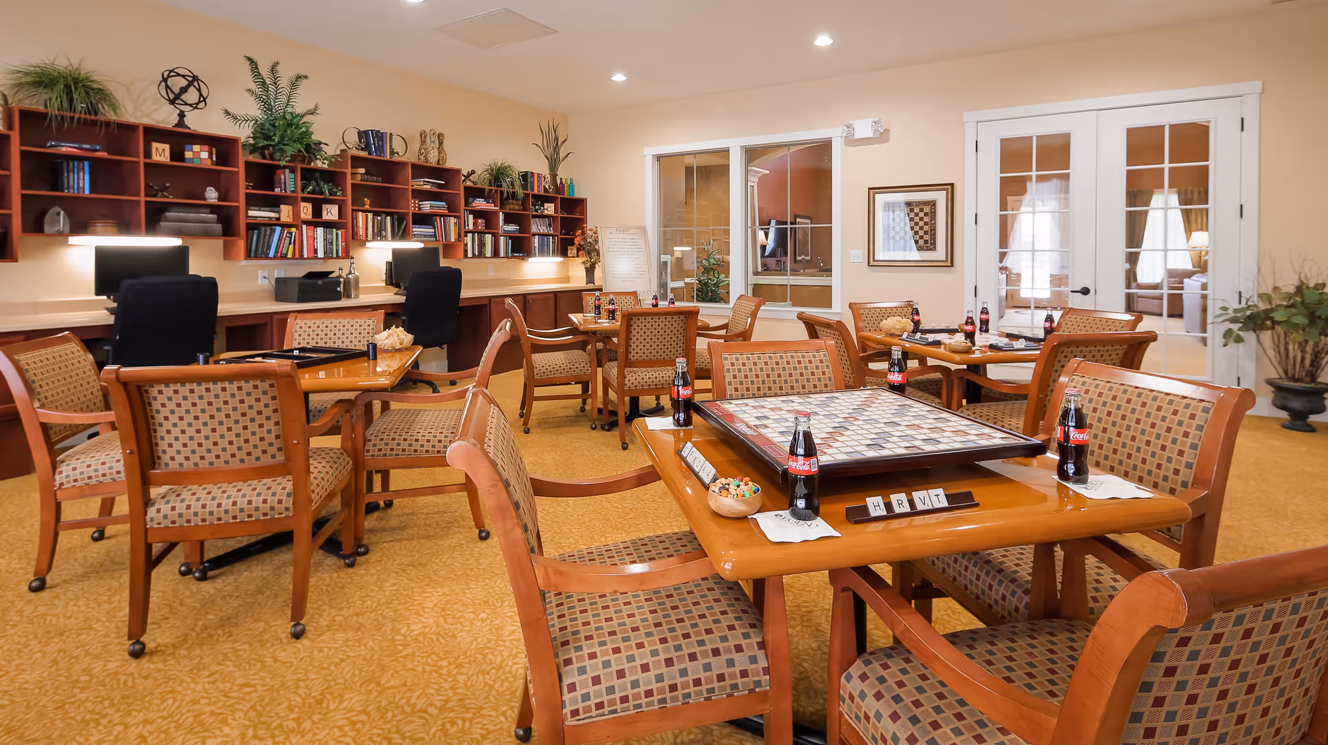 A well-lit activity room with multiple wooden tables and cushioned chairs arranged for board games. One table features a large checkerboard with game pieces and letter tiles. Several bottles of Coca-Cola are placed on the tables. In the background, there are desks with computers, office chairs, and shelves filled with books and decorative plants. The room has beige walls, carpeted flooring, and glass doors leading to another room.