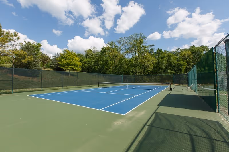 Outdoor tennis court with blue playing surface and green surrounding area, enclosed by a green fence. Trees and a partly cloudy sky are visible in the background.