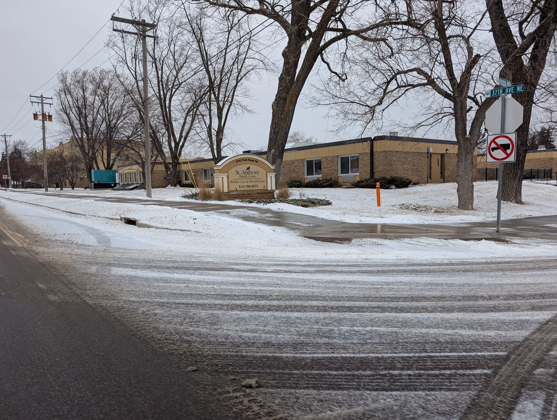 Exterior view of St. Anthony Health Center, a single-story brick building with leafless trees and snow-covered ground. A sign in front displays the facility name and contact number. Street signs indicate the intersection of Foss Road NE and 37th Ave NE. A no left turn traffic sign is also visible.
