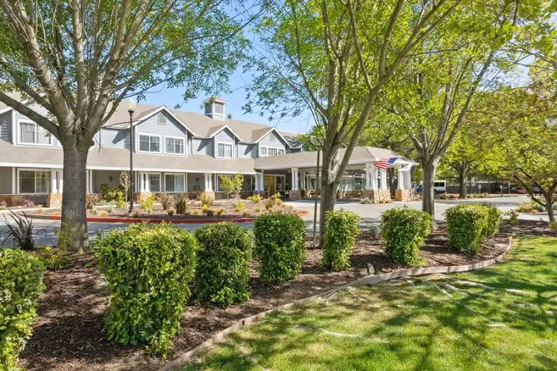 Exterior view of a senior living facility named Aegis of Carmichael with a well-maintained garden, green bushes, trees, and a driveway leading to the entrance. The building has a gray roof and multiple windows under a clear blue sky.
