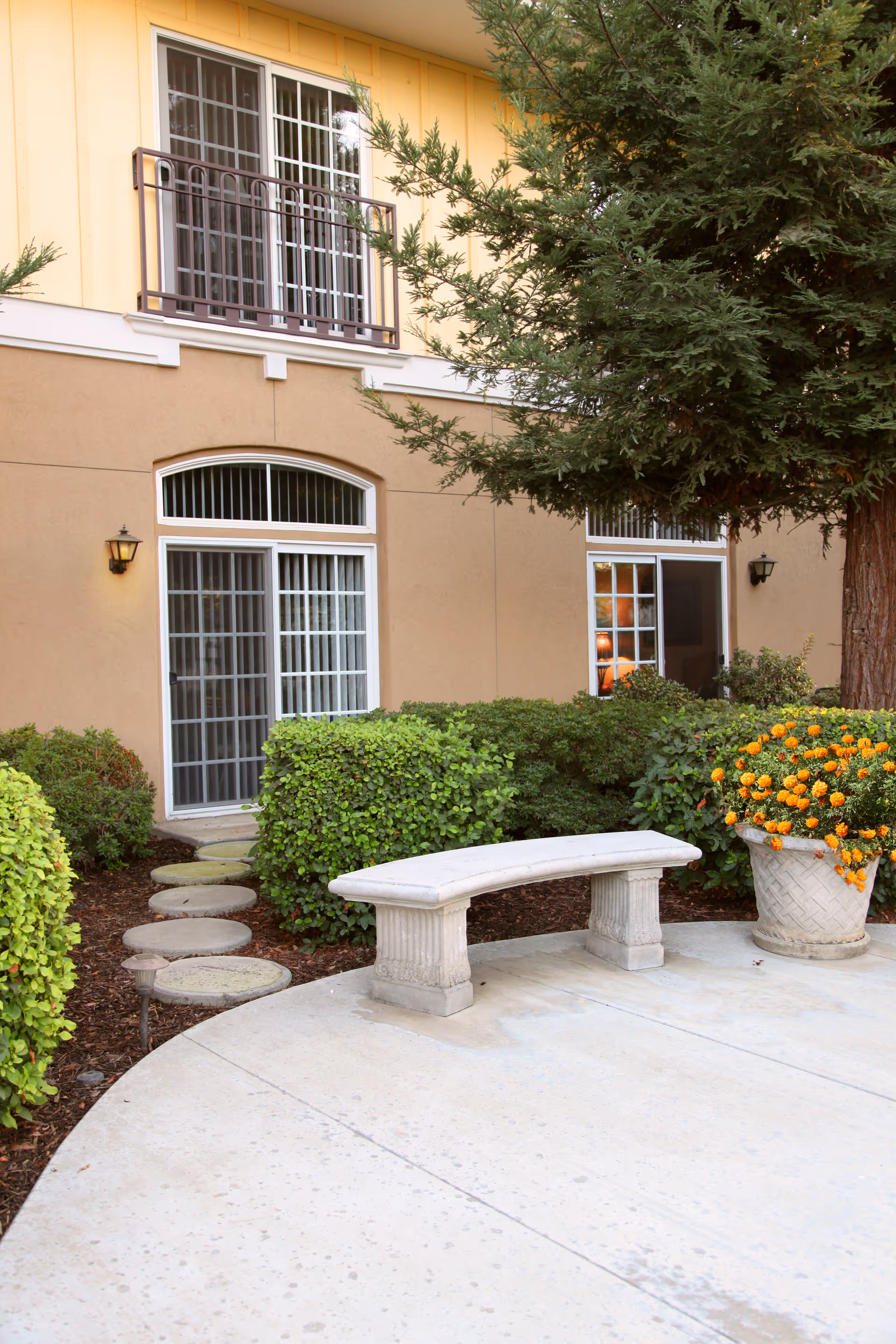 Outdoor patio area at Quail Park on Cypress featuring a curved stone bench, a large potted plant with orange flowers, green shrubs, a tall tree, and the exterior wall of the building with windows and sliding glass doors.