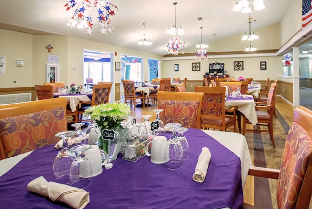 A brightly lit dining room with multiple tables covered in white and purple tablecloths. Each table is set with upside-down glasses, rolled napkins, and small flower arrangements. The chairs have patterned upholstery, and the room features hanging light fixtures decorated with red, white, and blue star-shaped ornaments. An American flag is visible on the right side of the room.