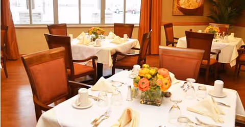 A dining room with several tables covered in white tablecloths, each set with folded napkins, cups, glasses, and silverware. There are wooden chairs with brown cushions around the tables, and a vase with colorful flowers is placed on the nearest table. Large windows with orange curtains allow natural light into the room.