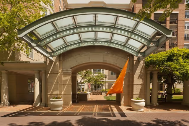 Entrance area of a senior living facility with a large glass and metal canopy over a brick-paved driveway. The building is made of light-colored bricks with multiple windows. There are two large white planters with flowers on either side of the entrance archway. An orange flag is hanging on the right side of the archway. Trees and greenery surround the entrance area.