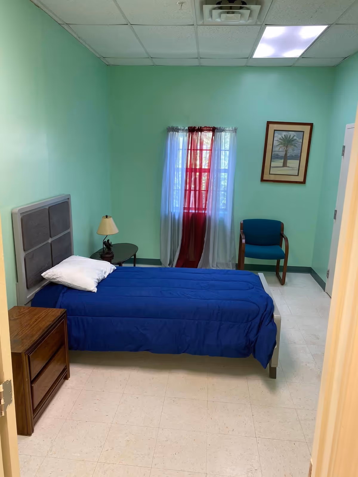 Small single-occupancy bedroom with a single bed covered in a blue comforter, a nightstand and lamp, a chair, mint-green walls, and a window with red curtains.