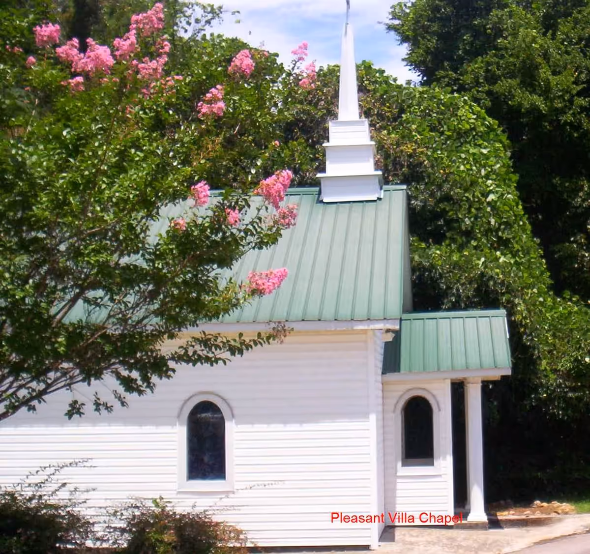 A small white chapel with a green metal roof and a white steeple, surrounded by green trees and pink flowering branches.