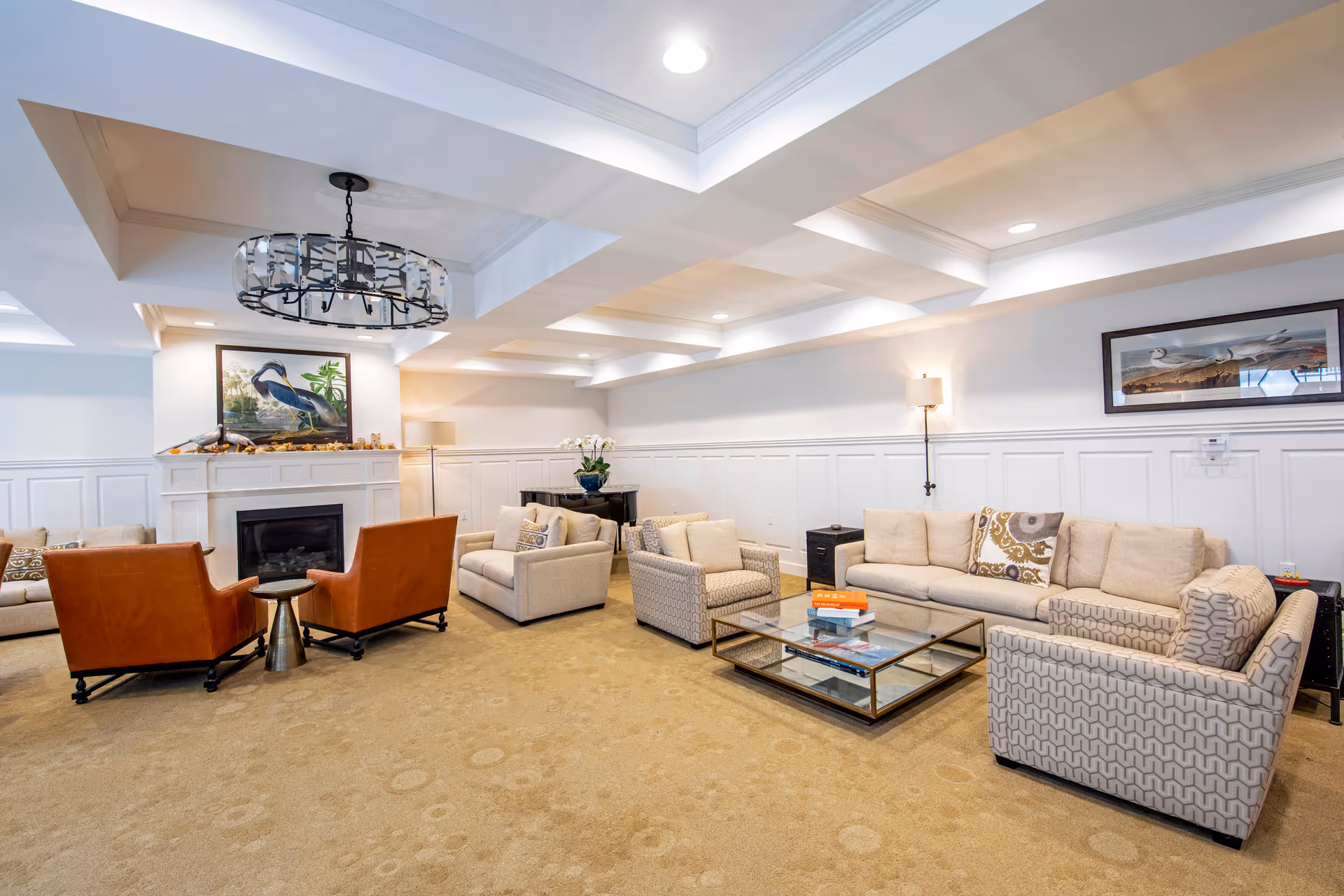 A spacious and well-lit living room with beige carpet and white paneled walls. The room features a large beige sofa, two patterned armchairs, two brown leather chairs, and a glass coffee table with books on it. There is a fireplace with decorative items and a painting of a bird above it. The ceiling has recessed lighting and a modern chandelier. Additional decor includes a floor lamp, a table with a flower vase, and framed artwork on the walls.