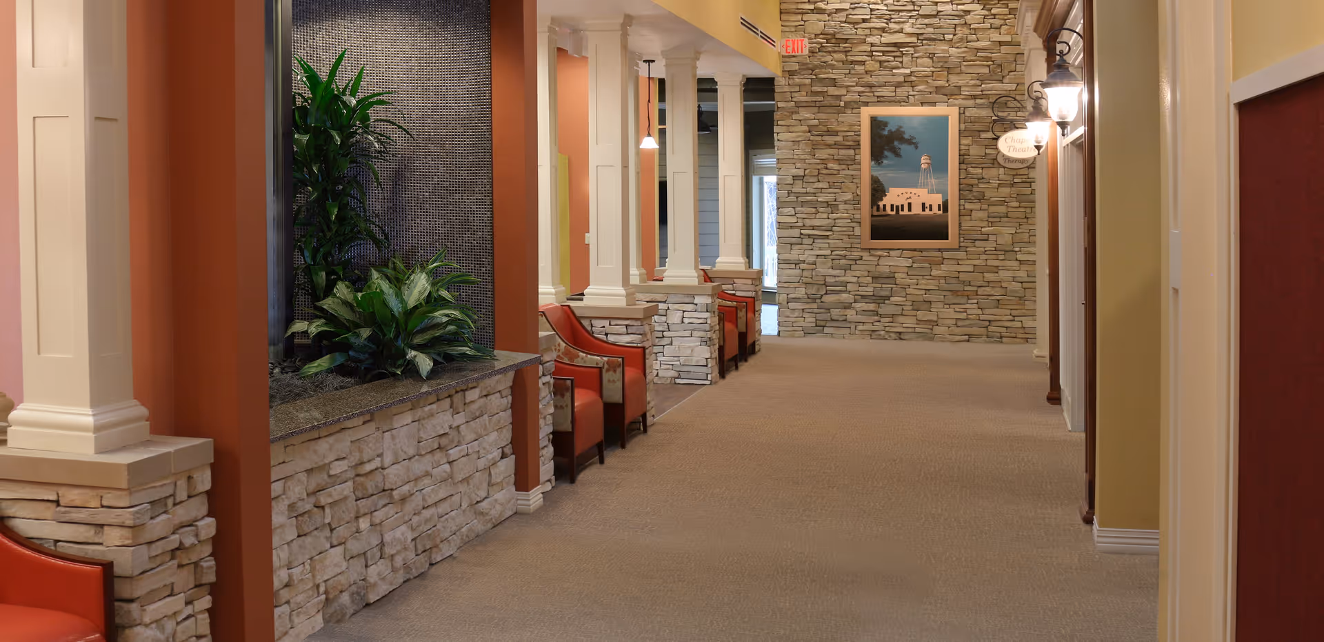 A wide hallway in a senior living facility with stone accent walls, beige carpet, and several red cushioned chairs along the left side. There are decorative columns and plants on the left, and a framed picture on the far wall. Warm lighting fixtures are mounted on the right wall.