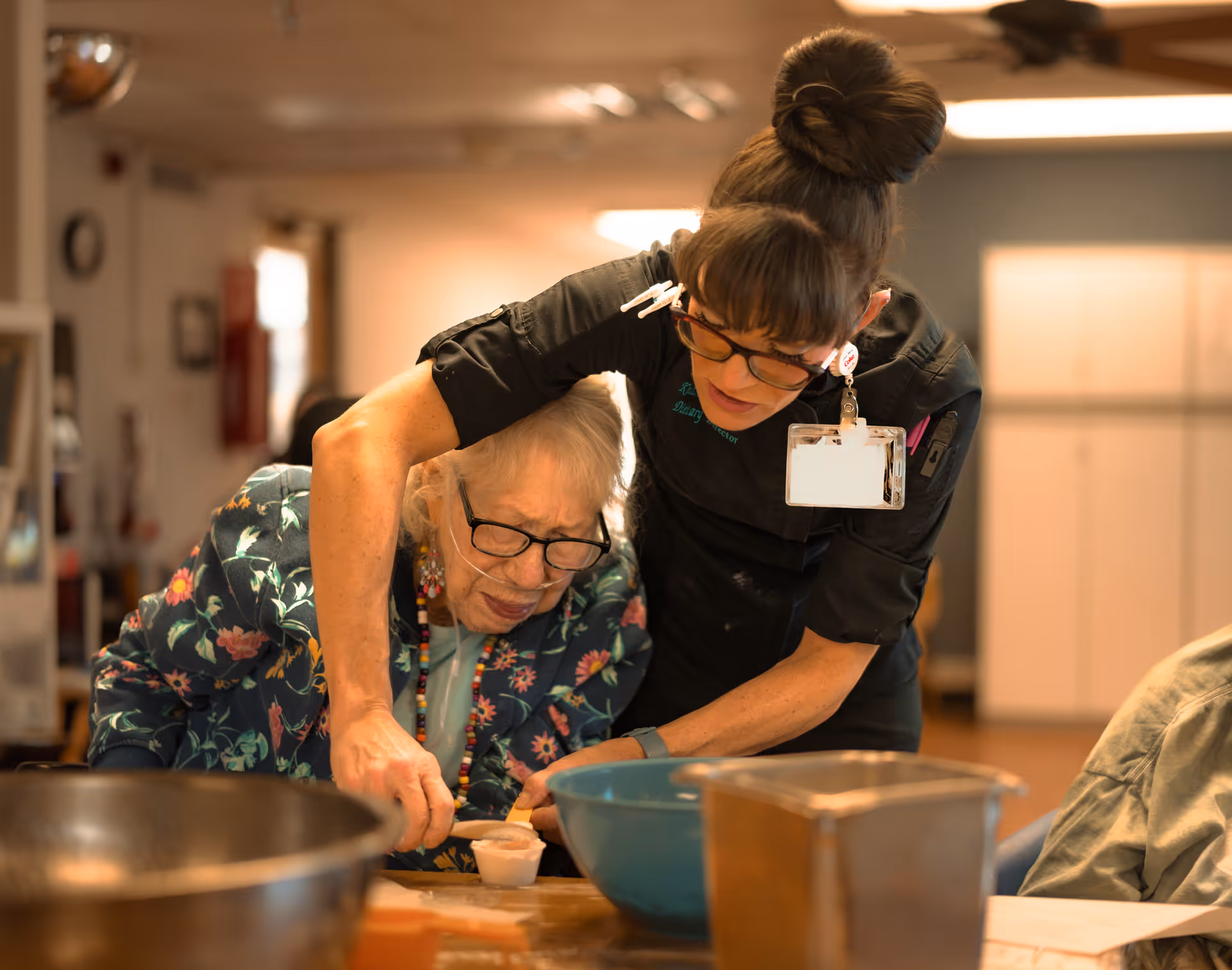 A caregiver assists an elderly woman with a cooking activity at a table. The elderly woman is wearing glasses and a floral patterned jacket, while the caregiver is leaning over her, guiding her hand. Various bowls and containers are on the table in front of them.