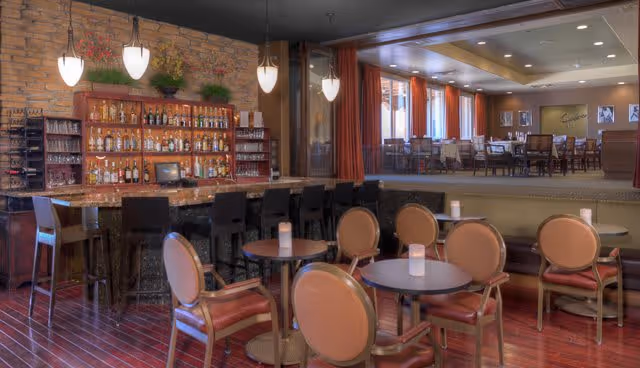 Interior view of a senior living facility lounge area featuring a bar with stools, shelves stocked with bottles and glasses, round tables with chairs, and a raised dining area with additional tables and chairs. The room has warm lighting, wooden floors, and large windows with curtains.