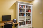 Small interior common area with a white bookshelf filled with books next to a desk with a computer monitor, phone, and chair against yellow walls.