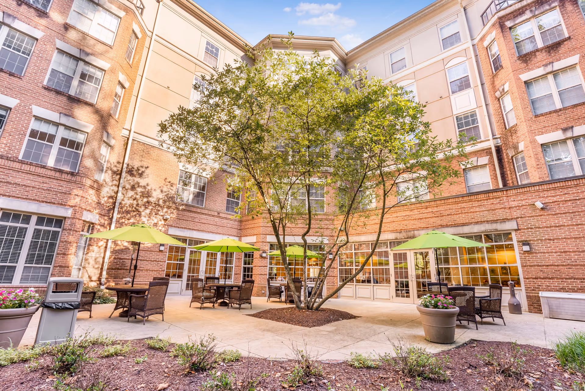 Outdoor courtyard area of a senior living facility with a large tree in the center, surrounded by patio tables with green umbrellas and wicker chairs. The courtyard is enclosed by a multi-story brick and beige building with many windows.