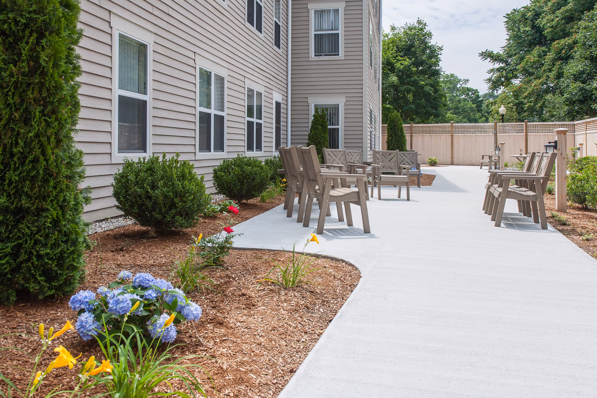 Outdoor patio area at The Residence at Pearl Street featuring a concrete walkway with multiple wooden chairs and benches arranged along the path. The patio is bordered by landscaped garden beds with green shrubs, blooming flowers including yellow daylilies and blue hydrangeas, and tall trees in the background. The building exterior is beige with multiple windows.