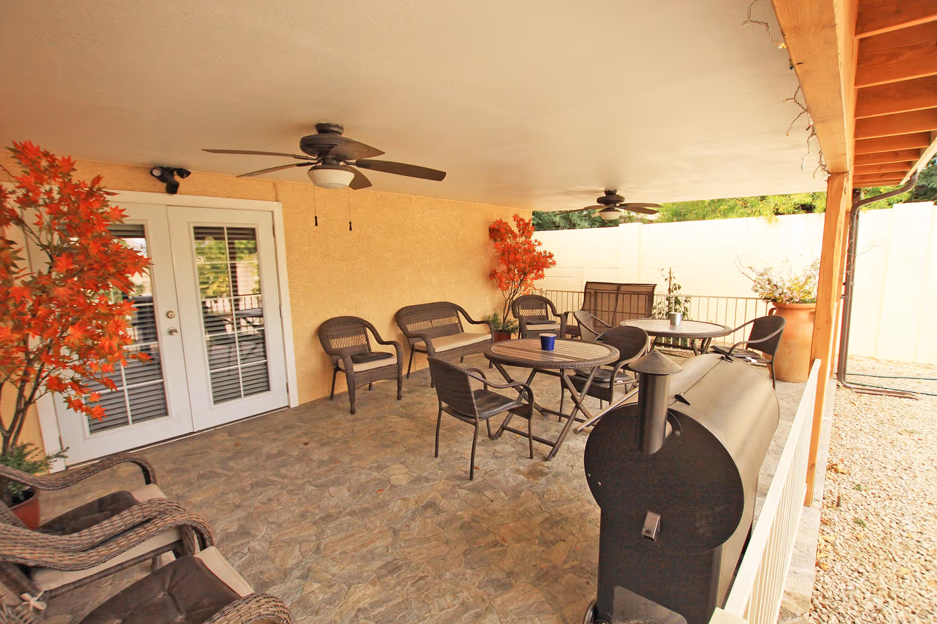 Covered outdoor patio area with stone flooring, several wicker chairs and tables, two ceiling fans, potted plants with red leaves, and a black barbecue smoker grill. The patio is enclosed by a white brick wall and has a wooden beam ceiling on one side.