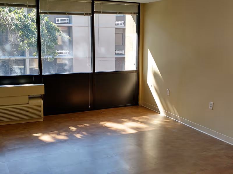 Empty room with large windows letting in sunlight, beige walls, and a tiled floor. There is a heating or air conditioning unit below the windows and electrical outlets on the walls.