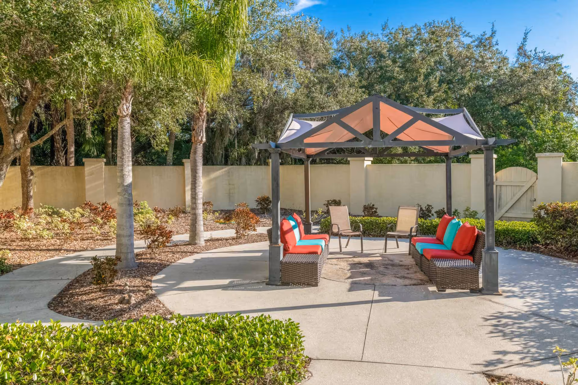 Outdoor seating area with a canopy structure providing shade over two wicker benches with red and turquoise cushions and two brown chairs, surrounded by trees, bushes, and a beige wall with a gate in the background under a clear blue sky.