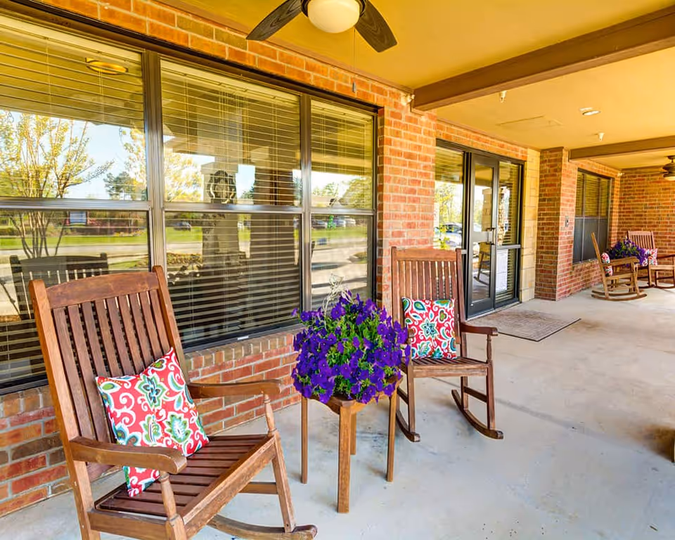 Covered entry porch with wooden rocking chairs, colorful pillows, and a planter of purple flowers in front of brick exterior windows and glass doors.