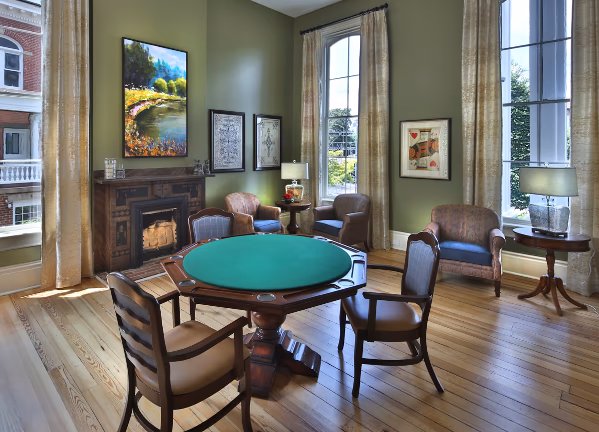Sunlit common sitting room with a round green-felt card table surrounded by wooden chairs, armchairs, a fireplace, large windows, and framed artwork.