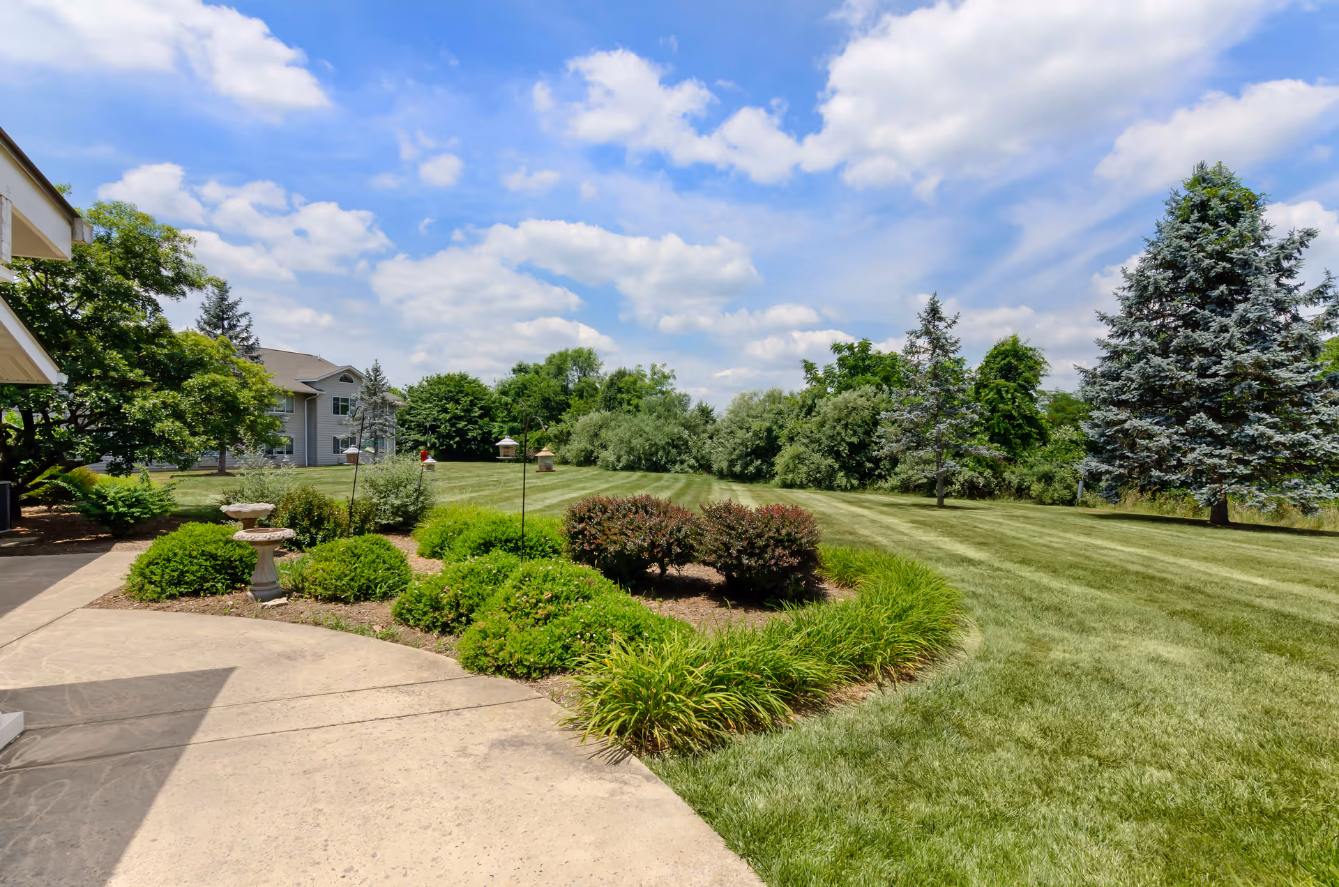 A well-maintained outdoor garden area with green grass, shrubs, and trees under a partly cloudy blue sky. A concrete pathway curves along the left side, and a building is partially visible on the left edge of the image.