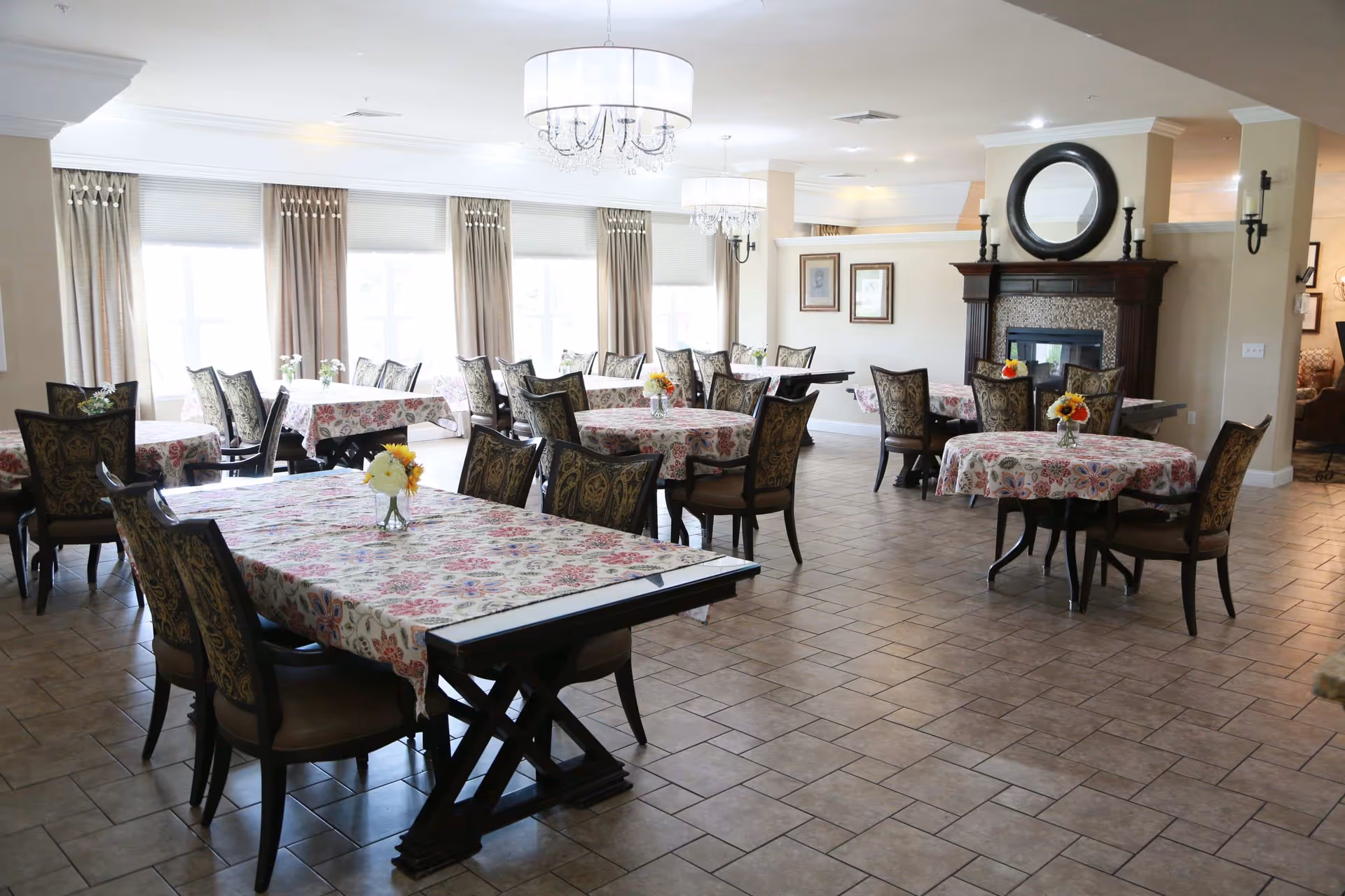 A bright dining room with several tables covered in floral tablecloths and decorated with small flower vases. The room features large windows with beige curtains, elegant chairs with patterned upholstery, tiled flooring, and a fireplace with a round mirror above it. Two chandeliers hang from the ceiling providing soft lighting.