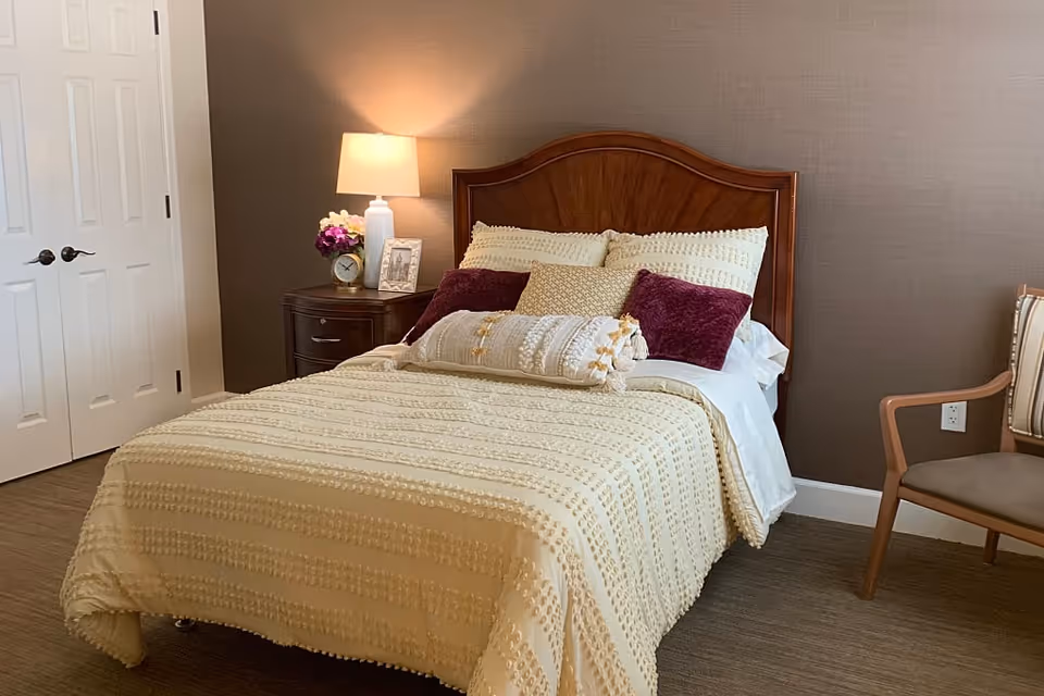 A cozy bedroom with a wooden headboard bed covered in a cream textured bedspread and multiple pillows in cream and burgundy colors. A nightstand with a lamp, a small clock, a framed photo, and a vase of flowers is beside the bed. There is a wooden chair with a cushioned seat and backrest to the right, and a closed white double door to the left.