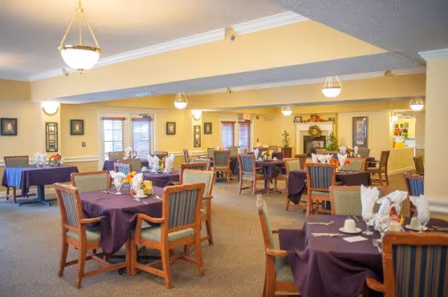 A spacious dining room with multiple tables covered in purple tablecloths, each set with white napkins, plates, and cups. Wooden chairs with green and striped upholstery surround the tables. The room has warm yellow walls, several windows with blinds, framed artwork, and ceiling lights providing illumination. A fireplace with decorations is visible in the background.