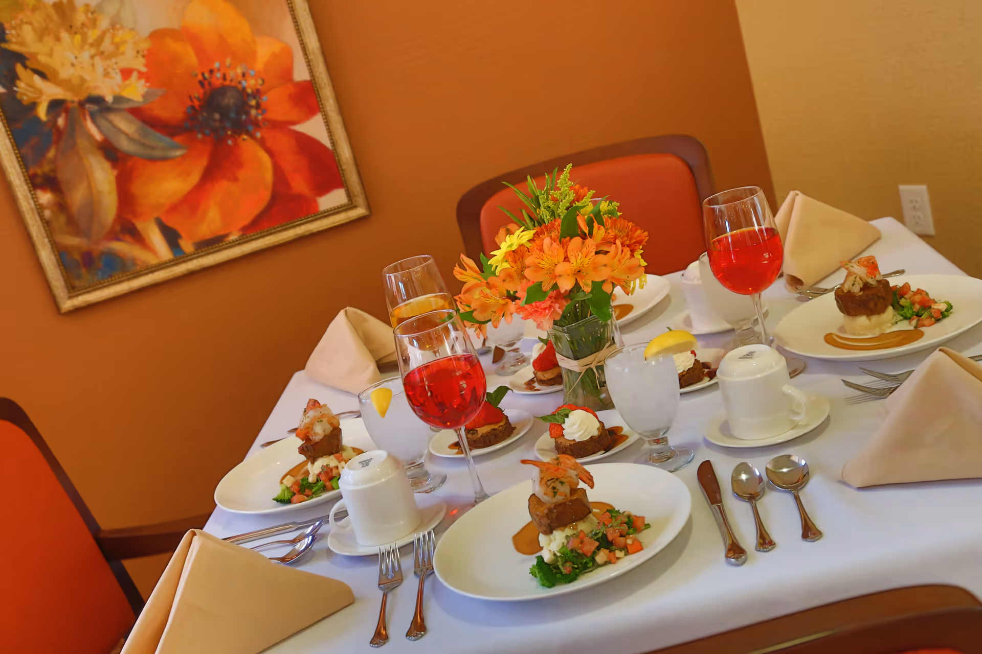 A dining table set for four with white tablecloth, beige napkins, silverware, and plates of gourmet food including shrimp and vegetables. There are glasses with red and clear beverages, a small vase with orange and yellow flowers, and a colorful floral painting on the orange wall in the background.