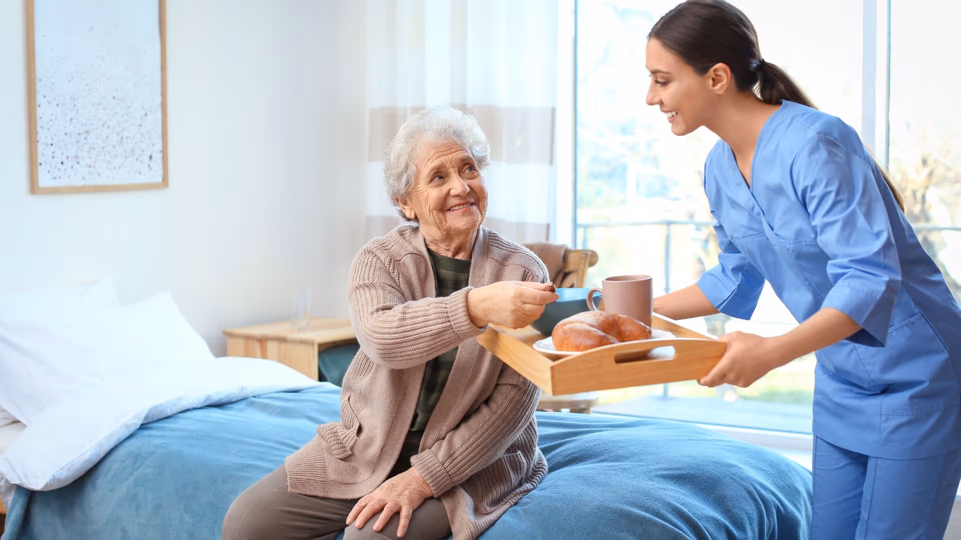 An elderly woman sitting on a bed in a cozy room, smiling and reaching out to take a cup from a young female caregiver dressed in blue scrubs who is holding a tray with a mug and croissants.