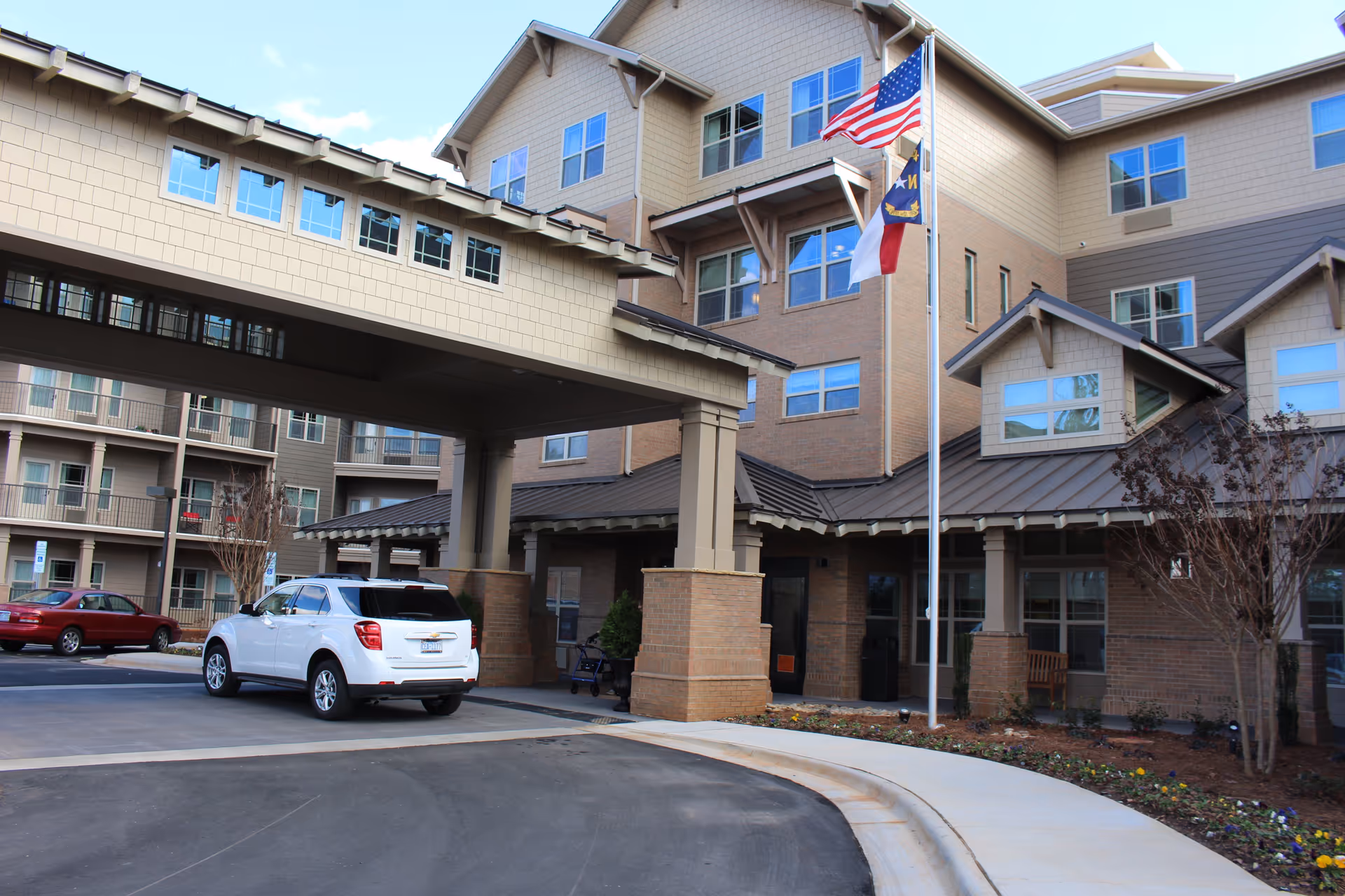 Exterior view of a multi-story senior living facility with a covered entrance. A white SUV is parked under the covered drop-off area. The building features multiple windows, brick and siding exterior, and a flagpole with three flags in front. There are landscaped flower beds and a curved driveway leading to the entrance.