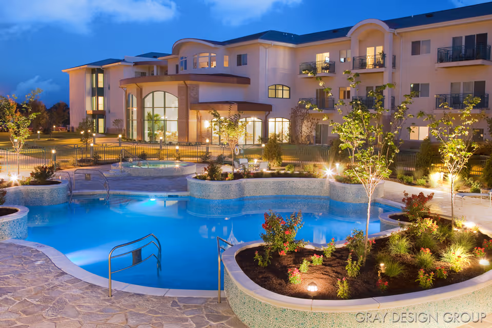 Evening view of a senior living facility with a well-lit outdoor swimming pool surrounded by landscaped garden beds and small trees. The multi-story building in the background has large windows and balconies, with warm interior lights visible.