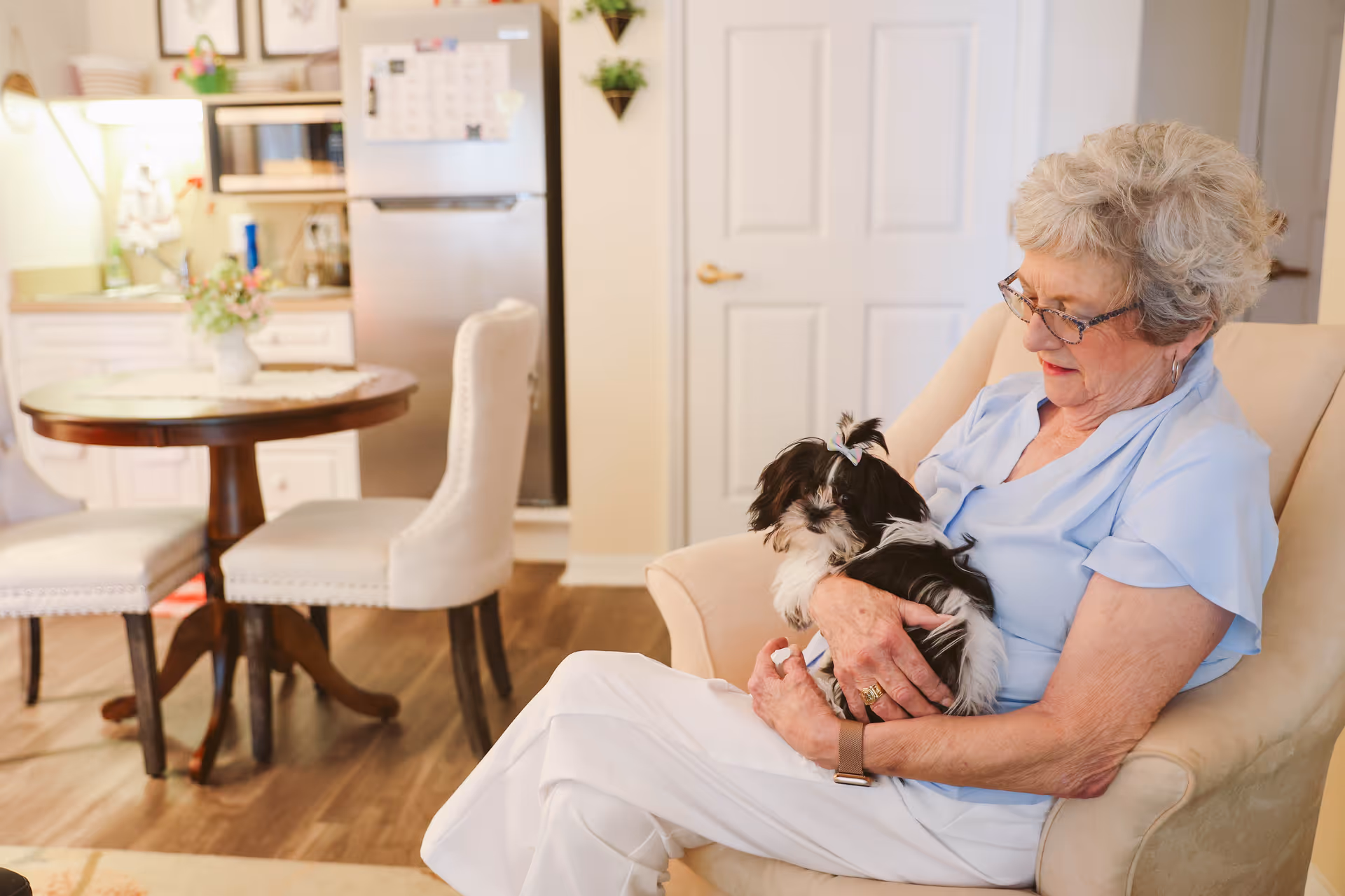 An elderly woman with short gray hair and glasses sits in a beige armchair holding a small black and white dog with a pink bow. Behind her is a small dining area with a round wooden table and two upholstered chairs, and a kitchen area with a refrigerator and cabinets.
