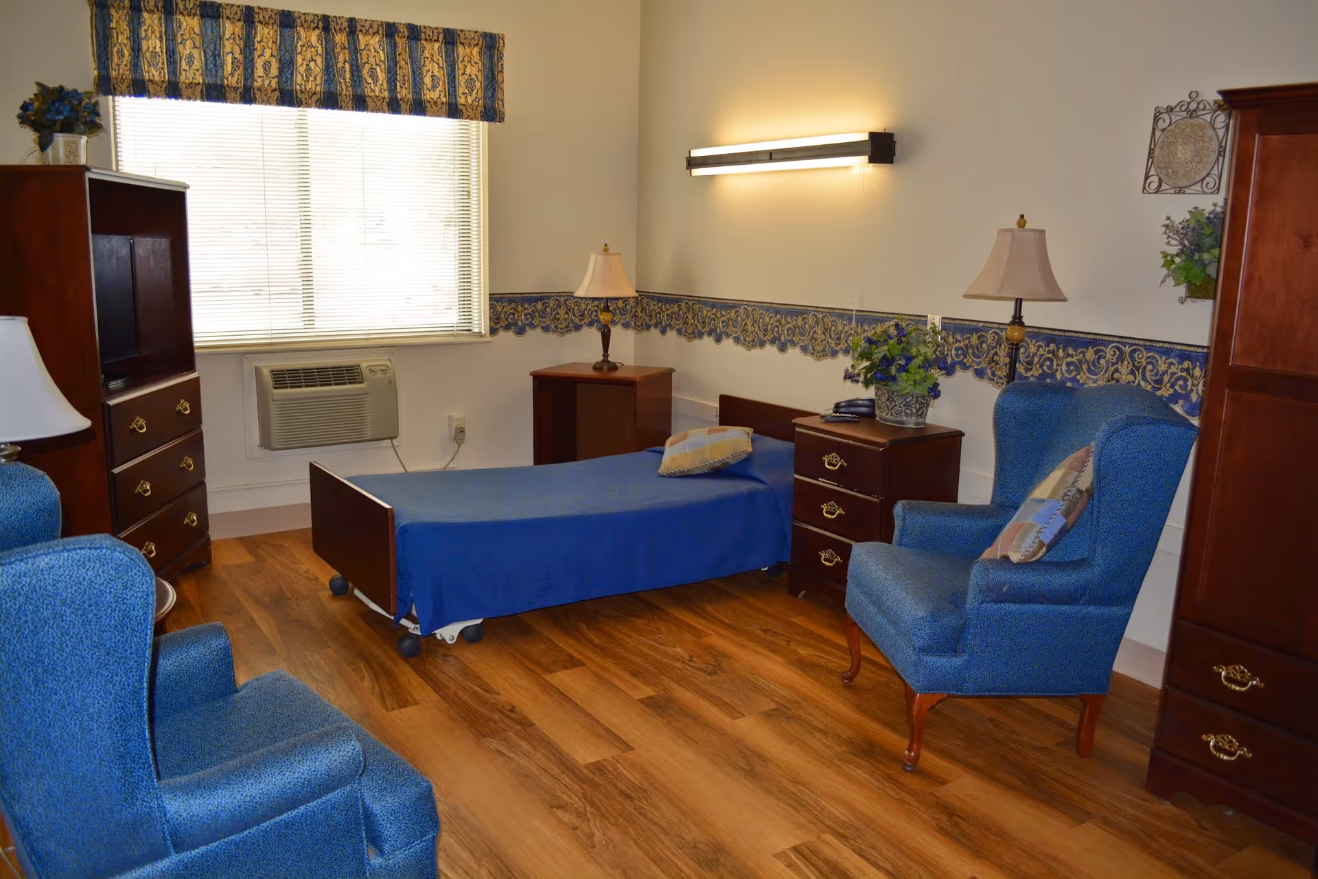 A cozy bedroom in a senior living facility featuring a single bed with a blue bedspread and a pillow, two blue upholstered armchairs with patterned cushions, wooden furniture including dressers and nightstands, table lamps, a window with blinds and a decorative valance, and a potted plant on a nightstand. The room has wooden flooring and a decorative wallpaper border along the walls.