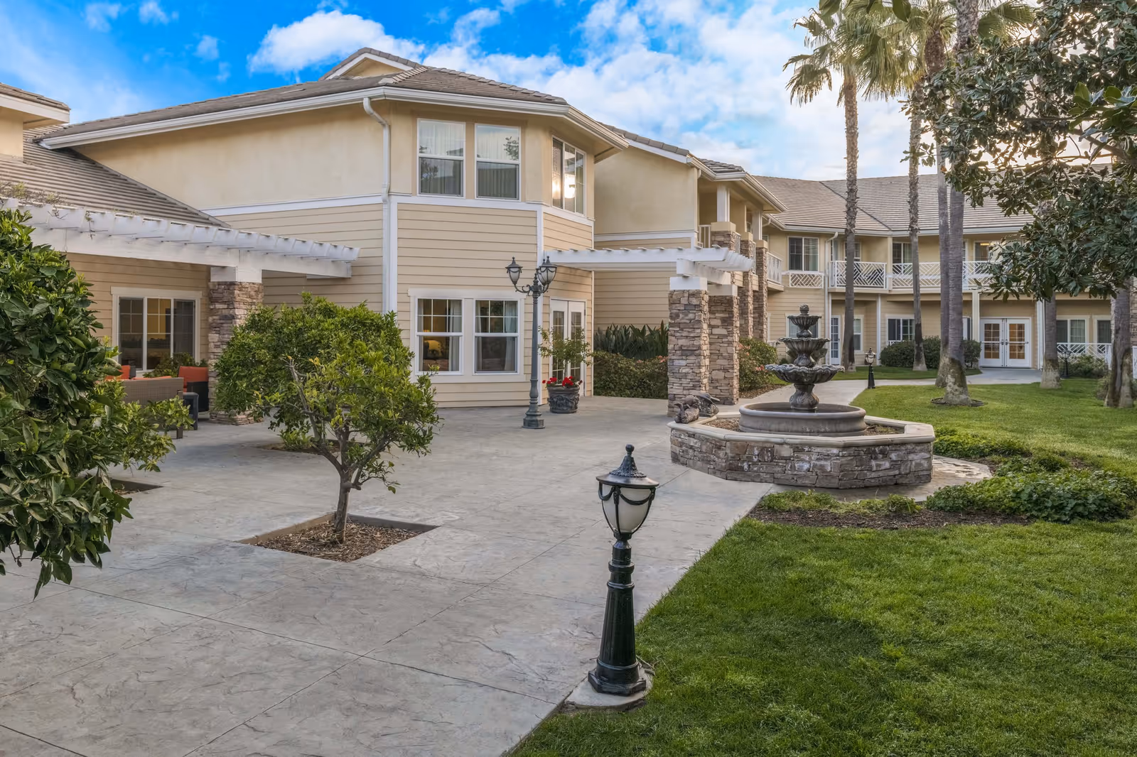 Courtyard with a stone fountain, lamp posts and landscaping in front of a two-story beige senior living building with palm trees.