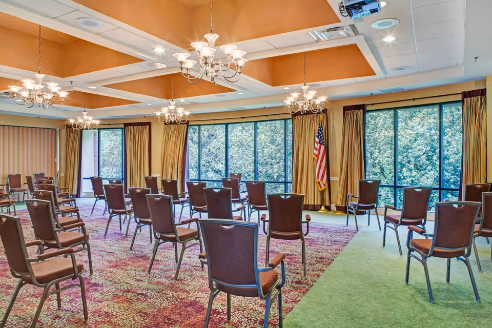 A spacious meeting or activity room with multiple brown chairs arranged in rows facing forward. The room has large windows with yellow and brown curtains, allowing natural light to enter and showing greenery outside. The ceiling features recessed lighting and several chandeliers. An American flag stands in the corner near the windows.