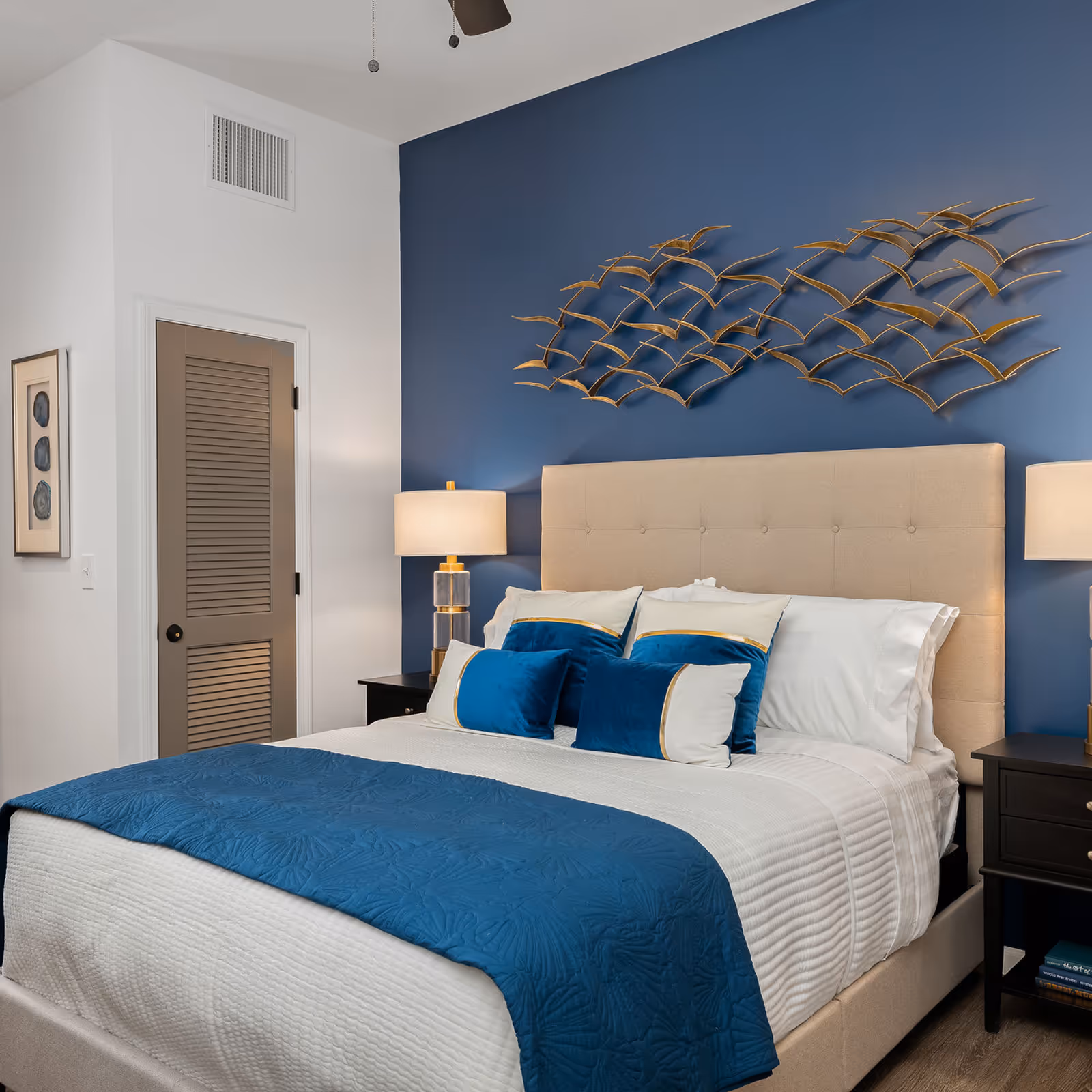 Well-decorated bedroom with a beige upholstered bed against a blue accent wall, blue bedding, decorative metal wall sculpture, and bedside lamps.