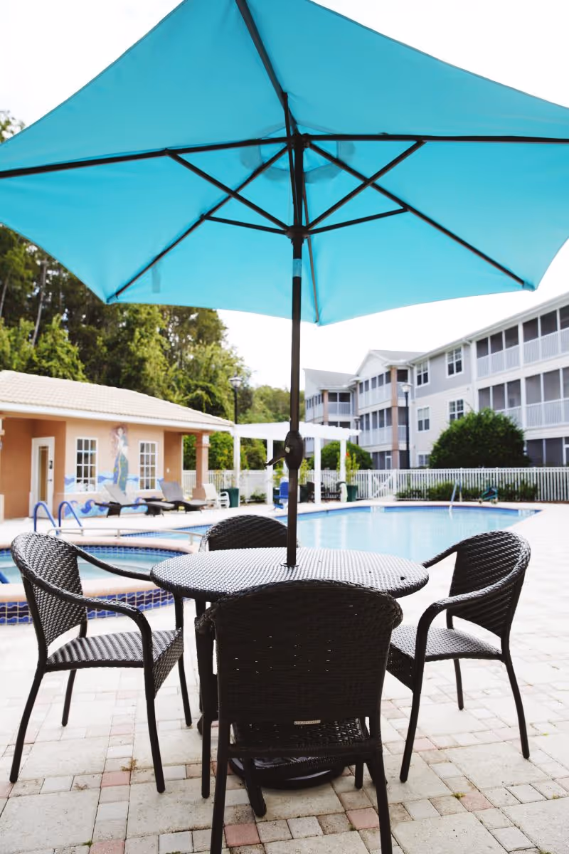 Outdoor seating area with a round table and four black wicker chairs under a large blue umbrella next to a swimming pool. In the background, there are lounge chairs, a small building with a mural, and a multi-story residential building surrounded by trees and a white fence.