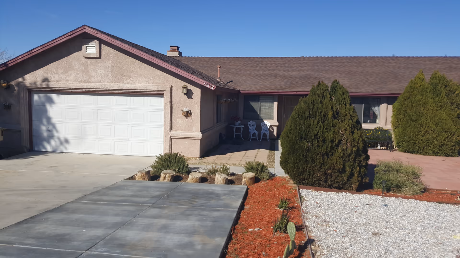 Front exterior view of a single-story house with a two-car garage, a concrete driveway, and landscaped front yard featuring bushes, red mulch, and white gravel under a clear blue sky.