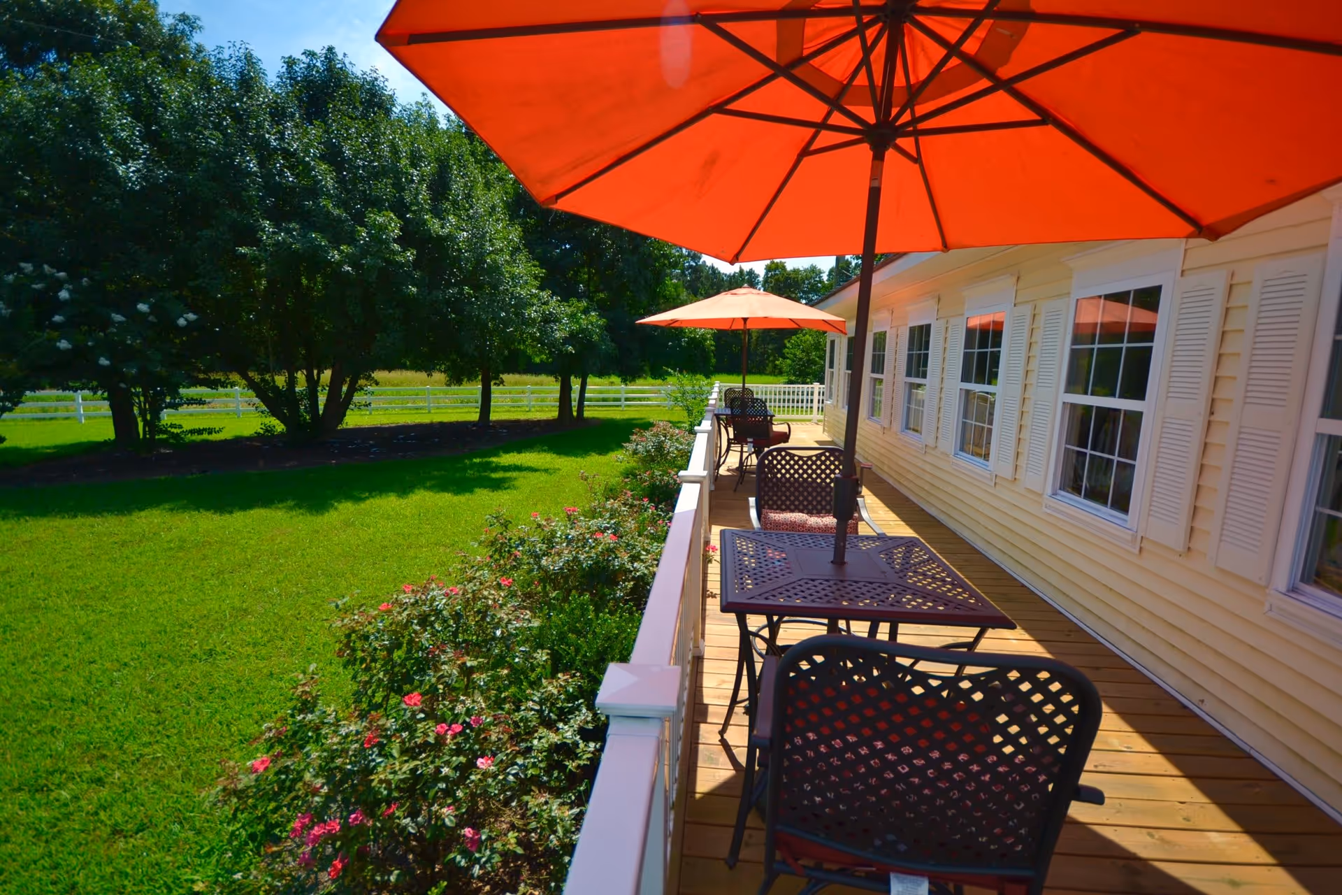 Outdoor patio area at Chesapeake Cottage Assisted Living with metal tables and chairs under large orange umbrellas, overlooking a green lawn with flowering bushes and trees in the background.