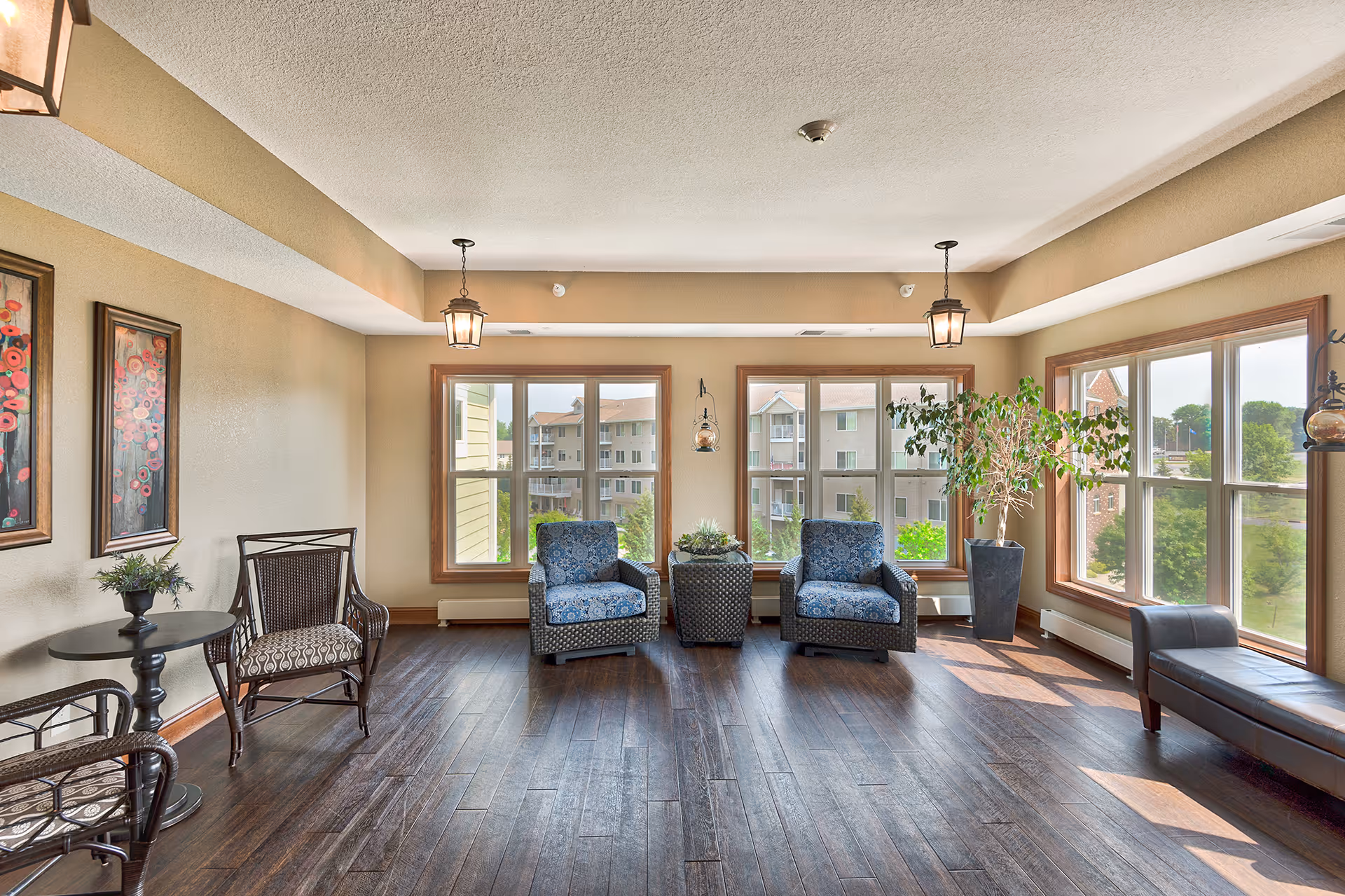A bright and spacious sitting area with large windows letting in natural light. The room features two blue patterned armchairs with a small table between them, a black leather bench near the windows, two wooden chairs with patterned cushions around a small round table, and a tall potted plant. The walls are beige with two framed floral paintings, and the floor is dark wood.